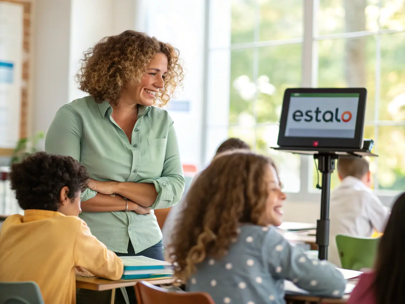A teacher smiling and interacting with students in a classroom setting, with a tablet displaying the Estalo platform interface in the background, showcasing the ease of integrating Estalo into daily teaching routines.